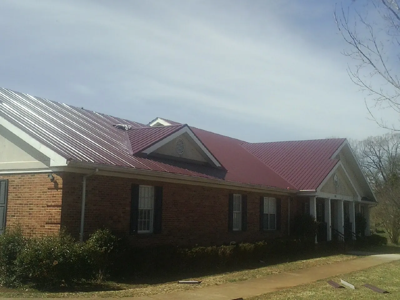 Red metal roof with gabled entries on a long brick building, from a Hail Damage Roof Repair job in Leo