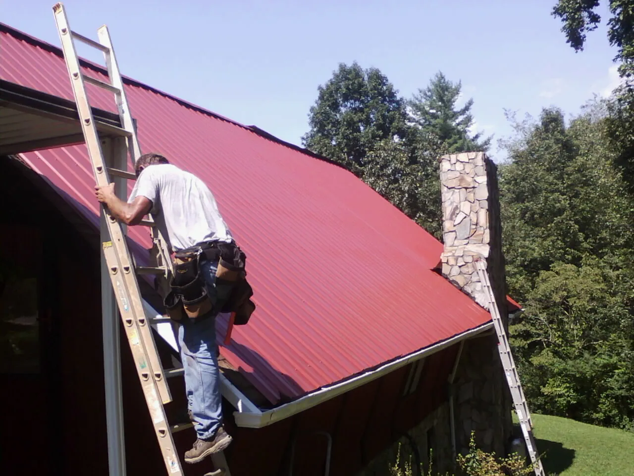 Roofer on a ladder beside a red metal roof and stone chimney — Soffit & Fascia Repair by Roofing Brothers in Leo